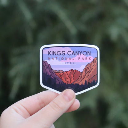 Fingers hold the Kings Canyon National Park sticker close, its purple sky and red cliffs bright against dark pines.