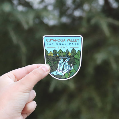 A thumb and finger hold the Cuyahoga Valley National Park sticker close, its waterfall art bright against dark pines.