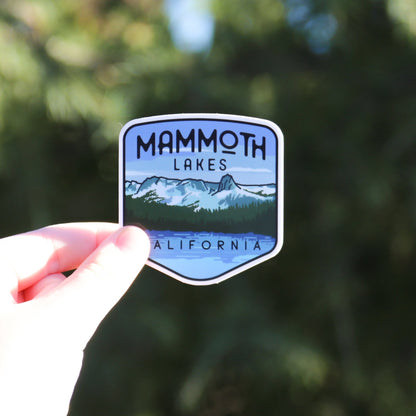 Fingers hold the Mammoth Lakes sticker in bright sun, its snowy peaks and lake bright against blurred pines.