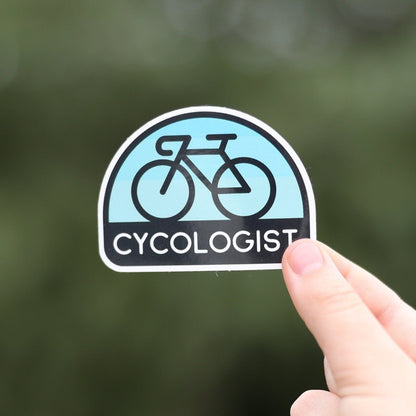 Held close to the camera against blurred green leaves, the Cycologist sticker shows its black bicycle on pale blue.