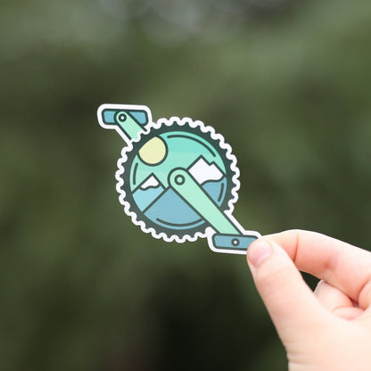 Fingers hold the Mountain Bike Crank sticker close to the lens, its snowy peaks and sun bright against blurred green.