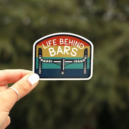 A hand with white nail polish holds the Life Behind Bars sticker close up against blurred evergreen branches.