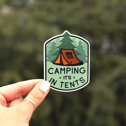 Held up against blurred evergreens, the Camping It's In Tents sticker shows an orange tent among pines.