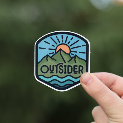 Fingers pinch the Outsider sticker close to the lens, its sunrise and mountain badge sharp against blurred green.