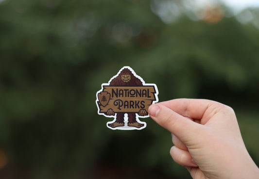 A hand lifts the Happy Bigfoot National Parks sticker toward a dense evergreen, the sticker small in frame.