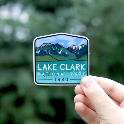 Fingers hold the Lake Clark National Park sticker against blurred green foliage, snowy peaks above a blue lake.
