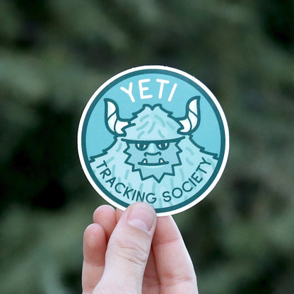 Fingers hold the round Yeti Tracking Society sticker close to the camera, its horned snow creature in teal.